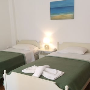 A hotel room with two single beds, each with a green bedspread and white pillows. There is a painting of a beach on the wall above the beds. A lamp is on the nightstand between the beds.