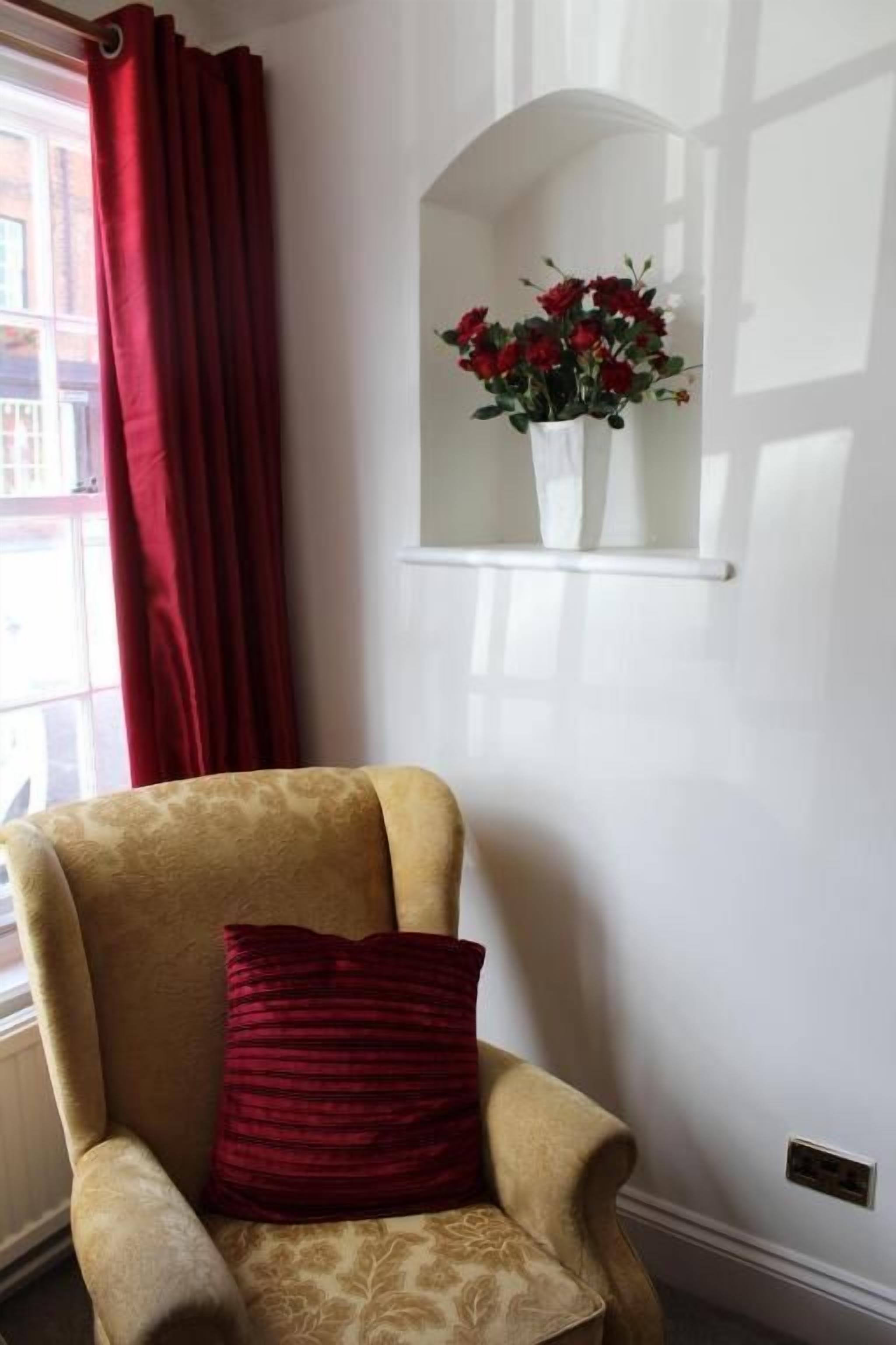 Une pièce avec un fauteuil beige, un coussin rouge, un mur blanc et une fenêtre avec des rideaux rouges.