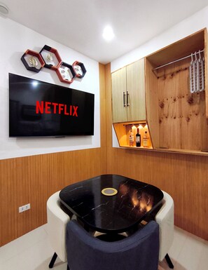 Une chambre moderne avec une table en marbre noir, un téléviseur fixé au mur affichant le logo de Netflix, des meubles en bois et une petite étagère avec des bouteilles.