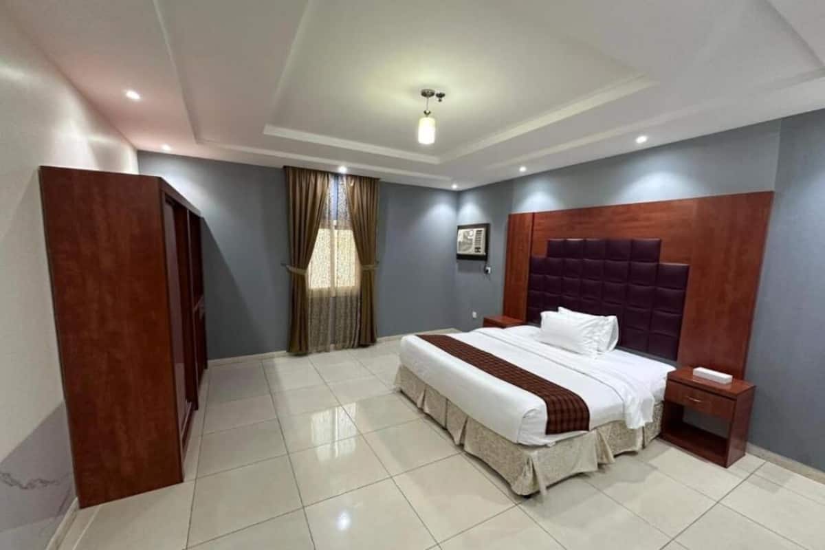A hotel room with a large bed, a nightstand, a wooden headboard, a ceiling light fixture, and a window with curtains.