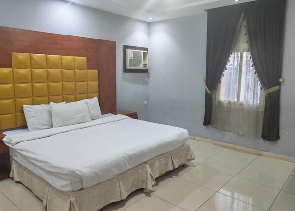 A hotel room with a large bed, a wooden headboard, an air conditioning unit, a window with curtains, and a tiled floor. - Furnished apartments (Jeddah)