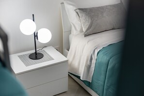 Ein ordentlich bezogenes Bett mit weißer Bettwäsche und einem Türkis-Akzent, ein Nachttisch mit einer modernen Lampe und ein weißer Beistelltisch.
