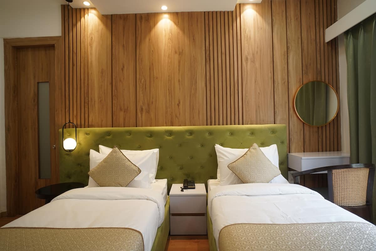 Una camera d'albergo con una testiera imbottita verde, due letti, un comodino con una lampada, uno specchio rotondo e una sedia in vimini.