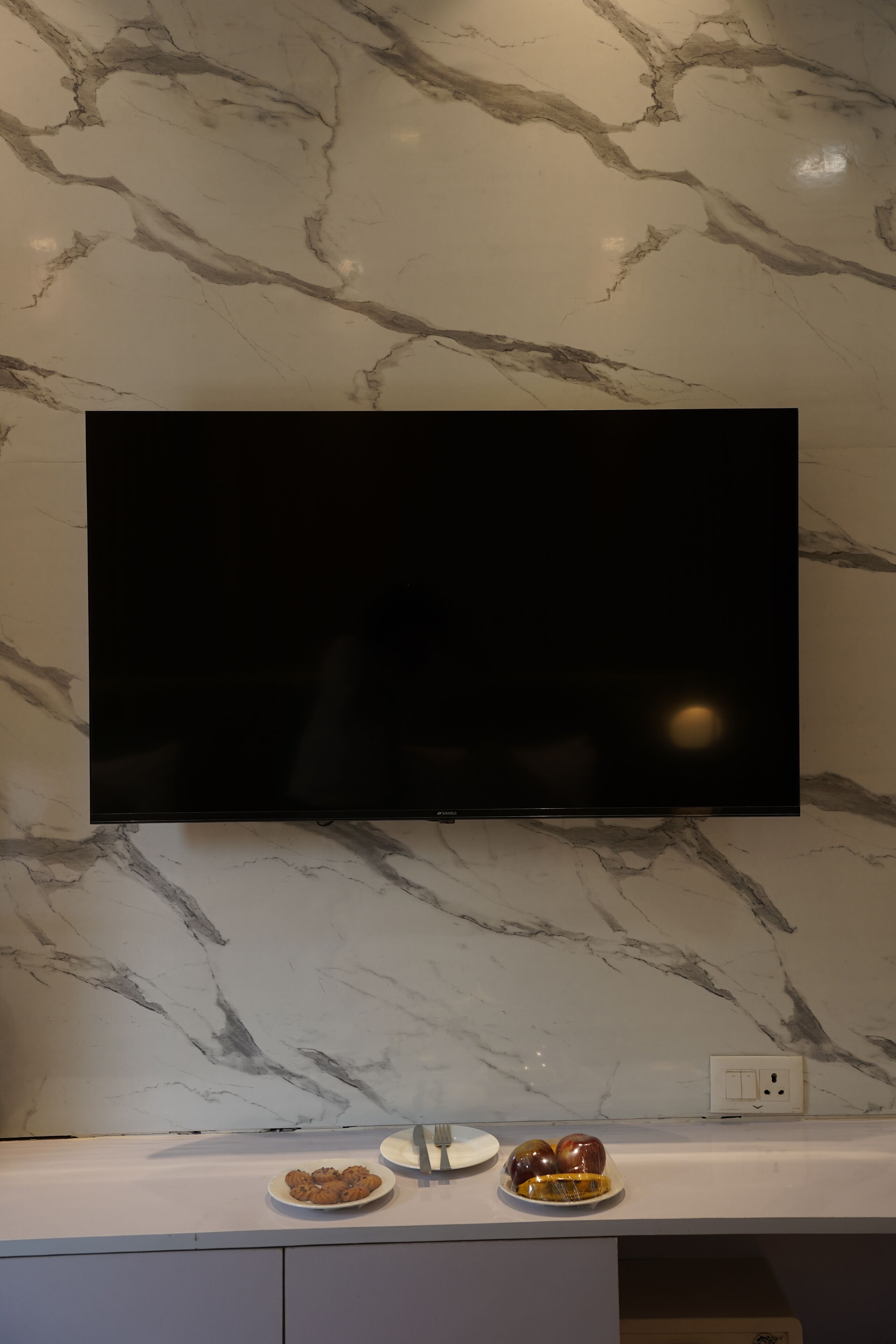 A flat-screen TV mounted on a marble wall, a white countertop with two plates of food, and a power outlet.