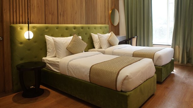Una habitación de hotel moderna con una cama tapizada en verde, paneles de madera en las paredes, un espejo redondo y un amplio ventanal con cortinas.