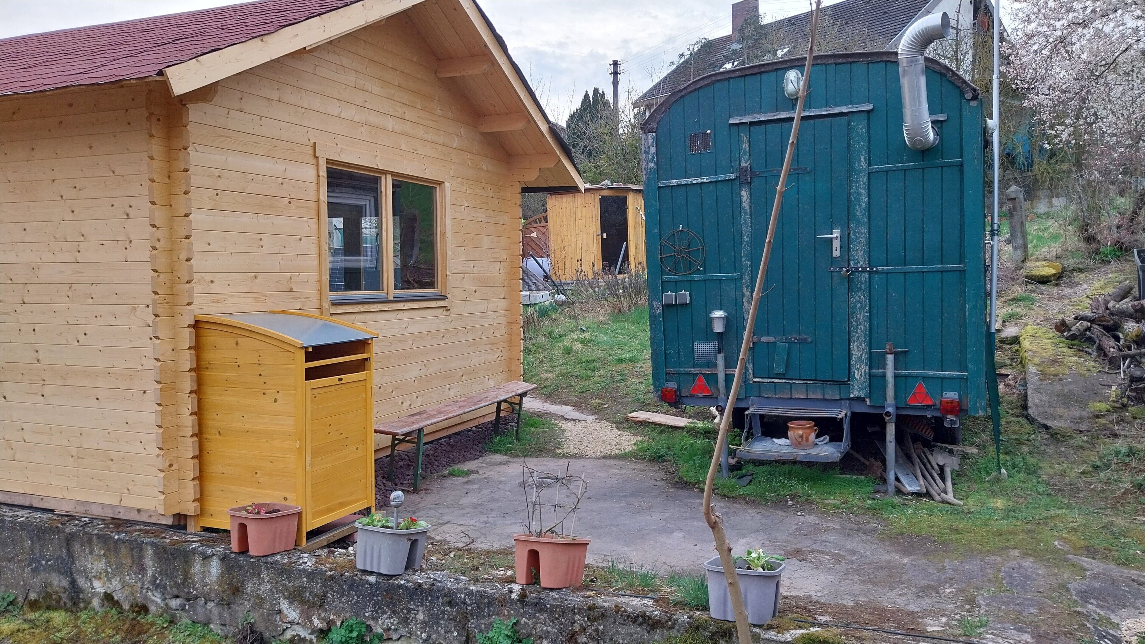 Pieni puutalo, jossa on pieni kuisti, vihreä vesisäiliö ja ruukuissa olevia kasveja.