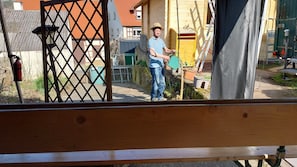 Eine Person steht neben einer Holzhütte mit einem Metallgitter, einer Leiter und einem grünen Gegenstand in der Hand.