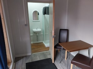 Une pièce avec un plancher en bois, une petite table avec une chaise, une porte donnant sur une salle de bain équipée d’un lavabo et d’un miroir, et un espace douche.