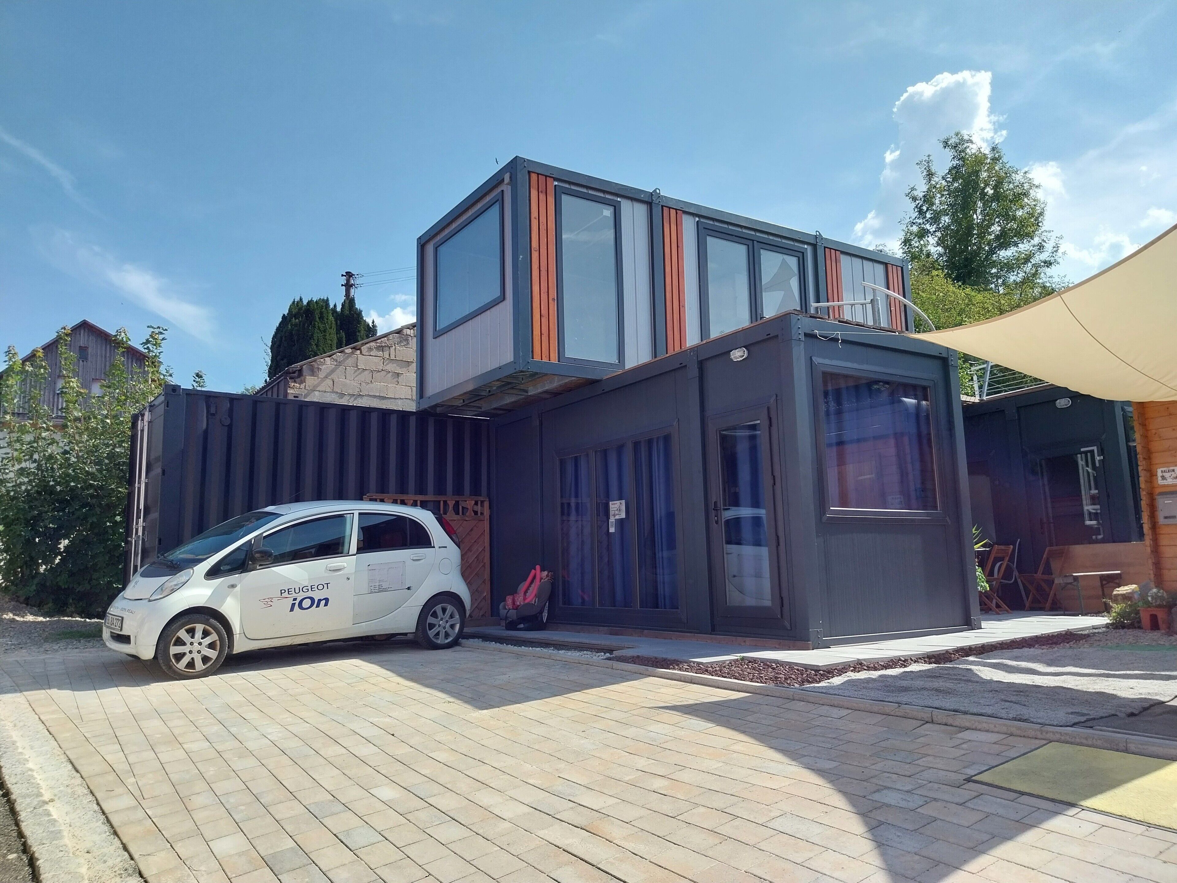 Un edificio moderno con un coche estacionado adelante, un pequeño espacio de terraza al aire libre y un cielo despejado.