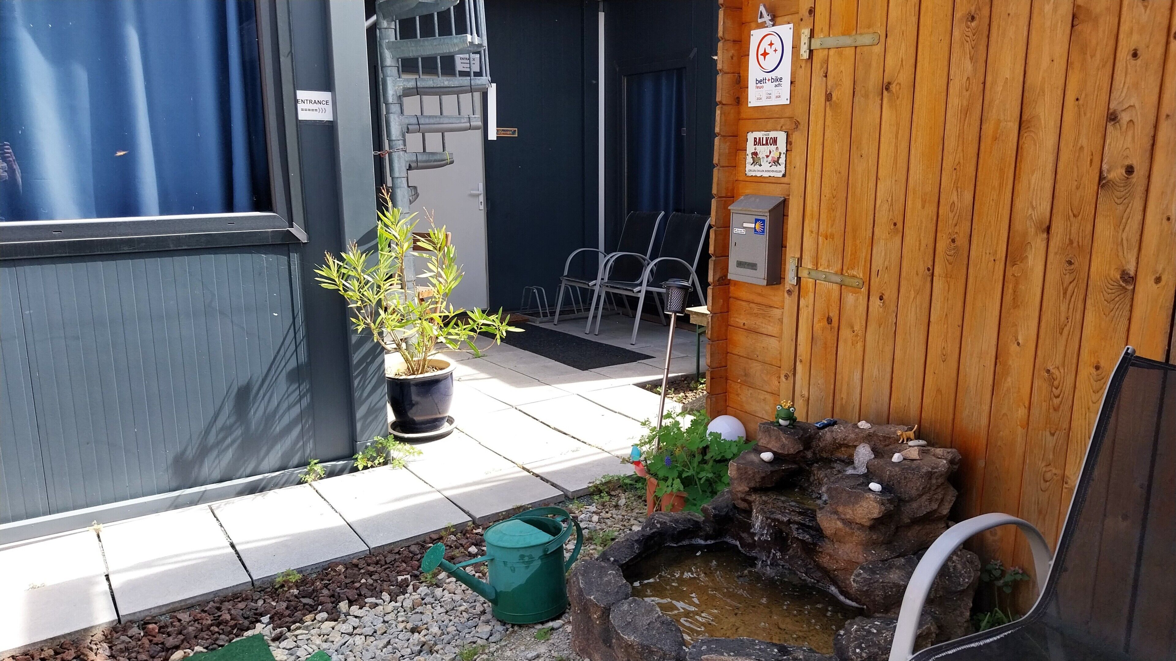 A small outdoor area with a wooden fence, a potted plant, a small garden with a watering can, and a sign indicating a balcony.