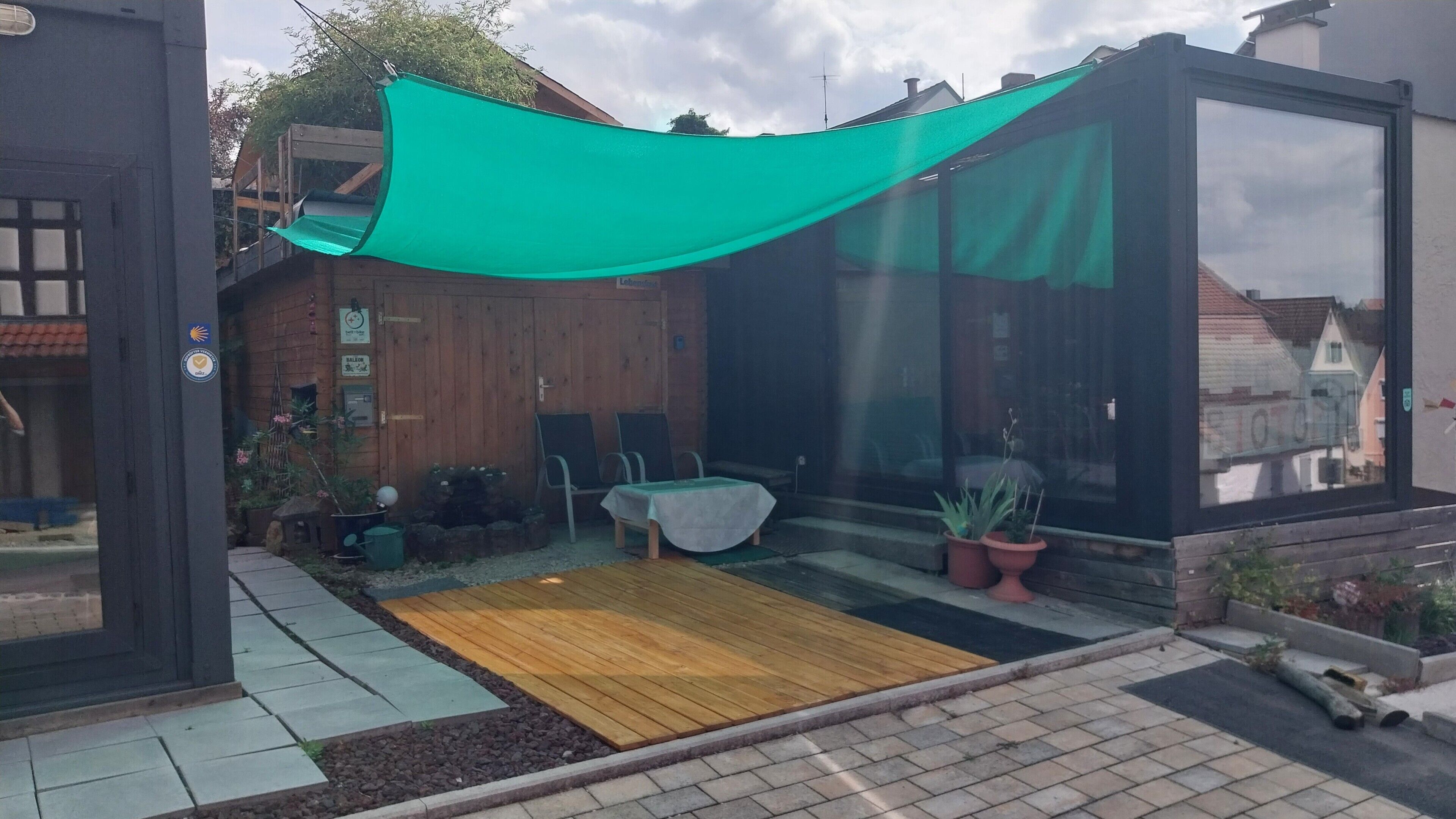 Eine Terrasse mit Holzdeck, Tisch und Stühlen, Topfpflanzen und einem grünen Sonnensegel.