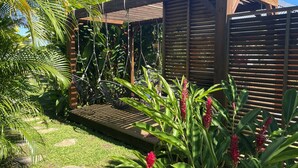 Eine Holzpergola mit Lamellen-Dach, umgeben von üppiger tropischer Vegetation und einem Kiesweg.