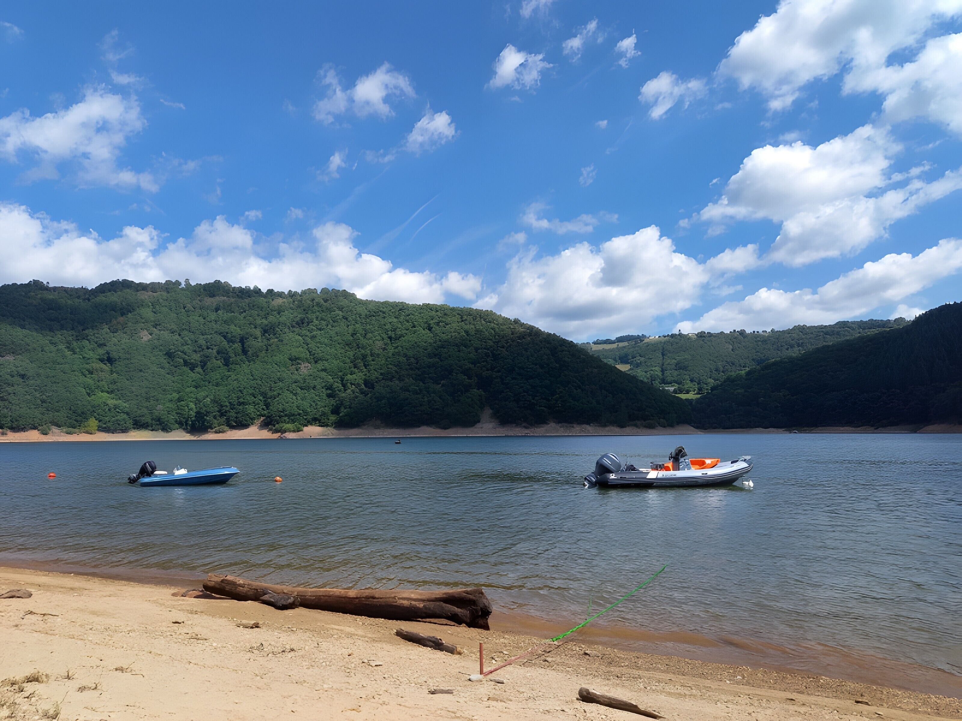 A calm lake with boats, a sandy beach, and forested hills in the background.