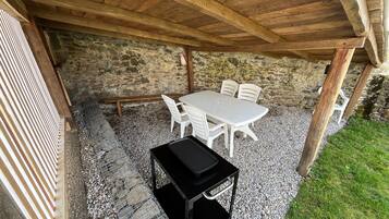 Eine überdachte Terrasse im Freien mit einer Steinwand, einem weißen Tisch und Stühlen sowie einem schwarzen Grill.