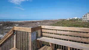 Um deck de madeira com um banco, com vista para uma praia e oceano, e edifícios ao fundo.