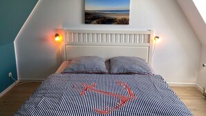 Ett sovrum med en säng, randiga sängkläder, en sänggavel, väggmonterade lampor och en tavla som föreställer en strandscen.