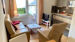 Ett vardagsrum med en soffa, en tv, ett litet bord och en köksdel med skåp och en mikrovågsugn.