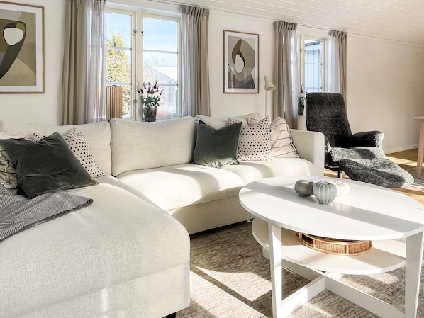 Un salon moderne avec un canapé d’angle, une table basse ronde et un fauteuil foncé. On y trouve deux fenêtres avec des rideaux, un vase de fleurs et des œuvres d’art abstraites accrochées au mur.
