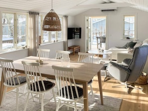 Un vaste salon comprenant une grande table à manger en bois, des chaises blanches, un fauteuil gris et un luminaire suspendu en osier.
