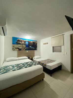 A bedroom with two beds, a large painting of a coastal landscape, a ceiling fan, and a window with blinds. - Hotel Suites 44 Laureles (Medellin)