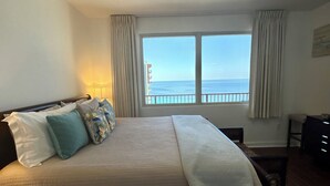 Quarto de hotel com uma cama, um ventilador de teto, vista para o mar e uma escrivaninha com uma cadeira.