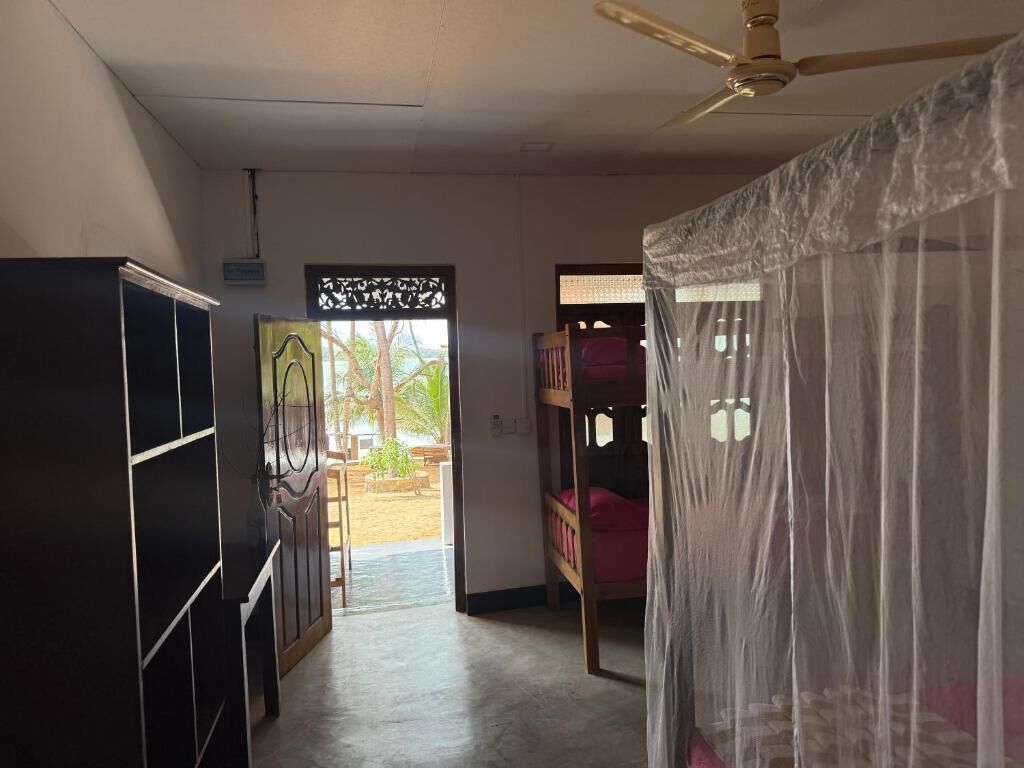 A room with a bed under a mosquito net, a ceiling fan, and a view of an outdoor area with a wooden bench and greenery.
