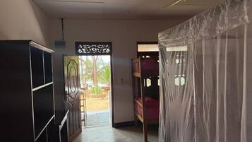 A room with a bed under a mosquito net, a ceiling fan, and a view of an outdoor area with a wooden bench and greenery.