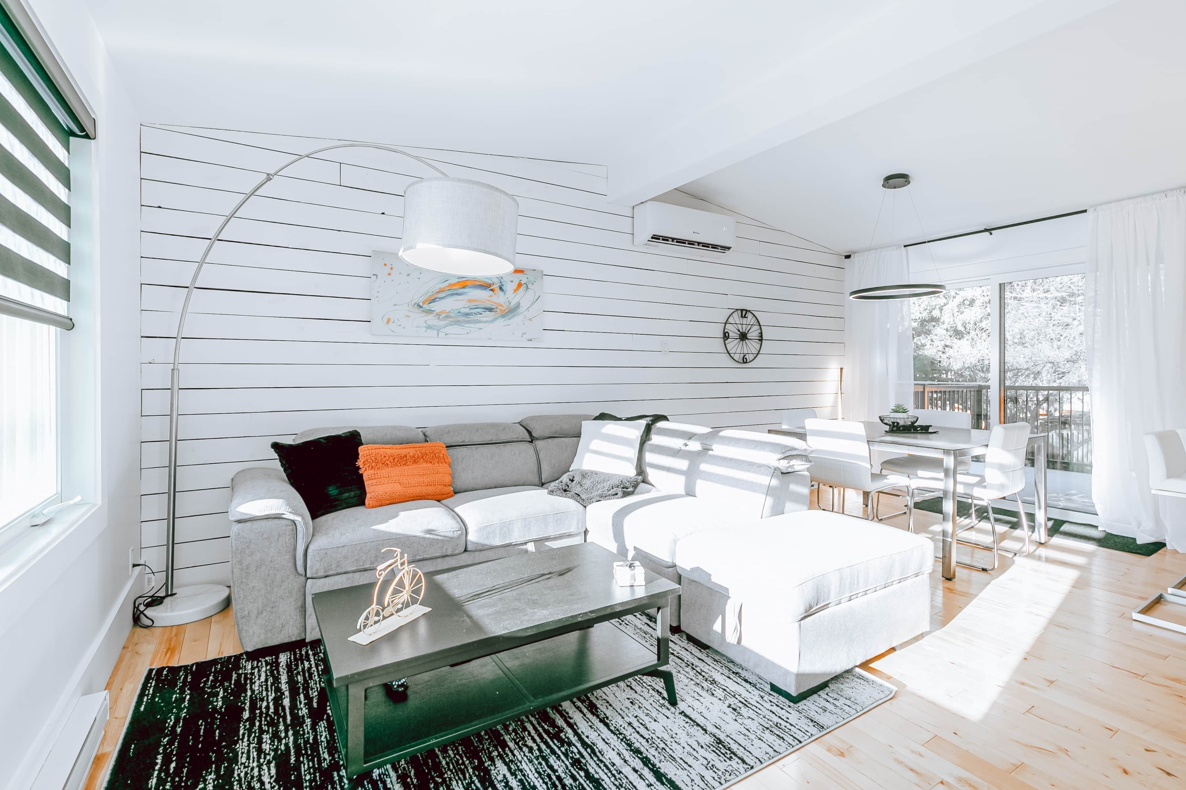 A modern living room with a grey sofa, white ottoman, and a glass coffee table. There is a dining area with a white table and chairs, and a large window providing natural light.