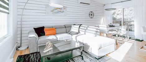 A modern living room with a grey sofa, white ottoman, and a glass coffee table. There is a dining area with a white table and chairs, and a large window providing natural light.