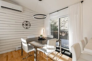Un coin repas avec une table en bois, des chaises blanches, une horloge et une vue dégagée sur l’extérieur grâce à une grande fenêtre.