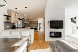 Une cuisine moderne dotée d’un îlot central, d’un coin repas avec une longue table, d’un téléviseur encastré et d’une cheminée.