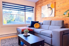A modern living room with a grey sofa, a wooden wall, a large window with blinds, a coffee table with a plant, and a painting. - Le Champery Tremblant 4 (Mont-Tremblant)