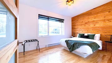 A bedroom with a wooden floor, a bed with white bedding, a window with blinds, and a nightstand with a lamp.
