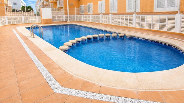 A swimming pool with a tiled deck, a white fence, and a building with balconies in the background. - Sun and Beach in La Mata (Torrevieja)