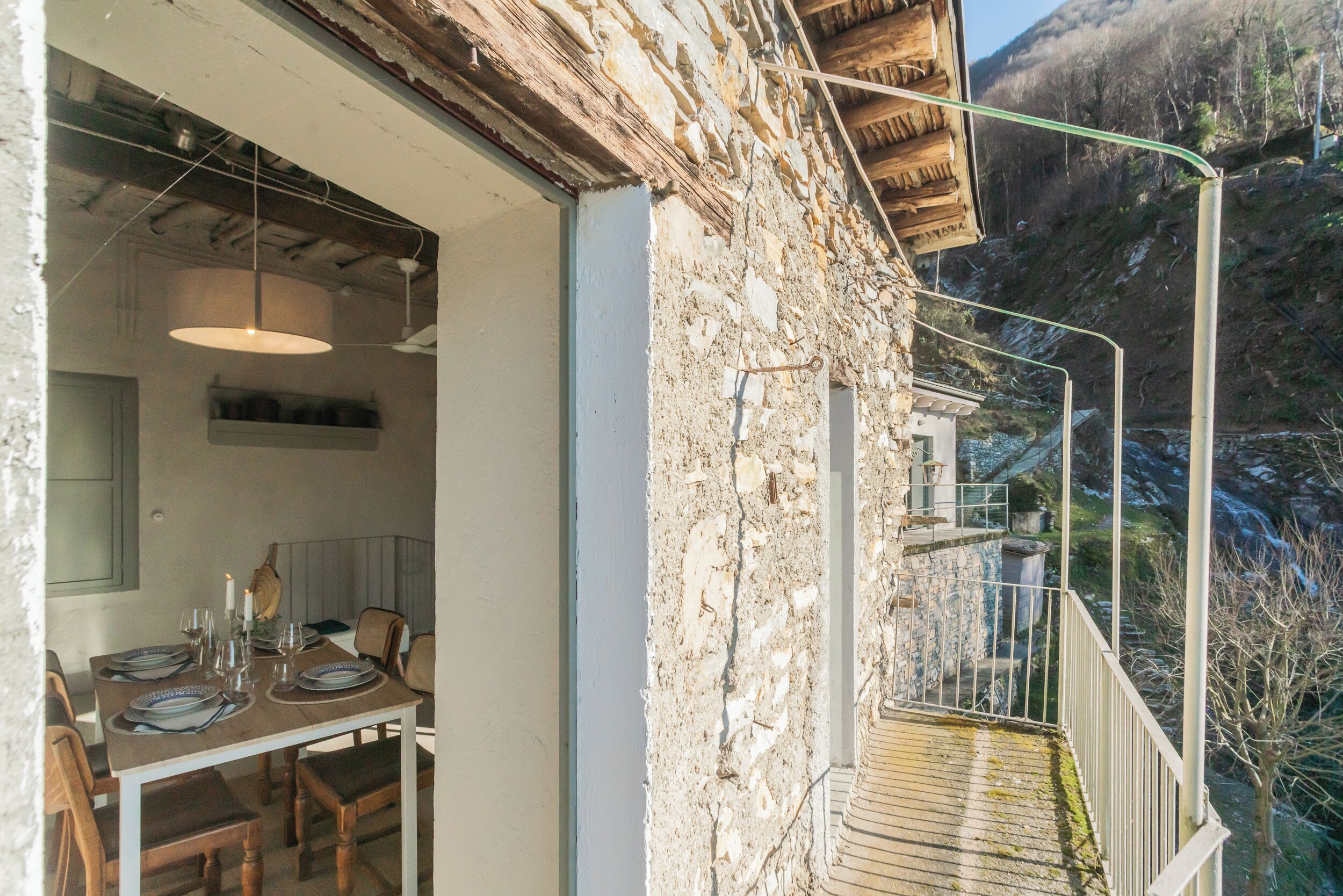 A rustic stone building with a dining area featuring a wooden table and chairs, a hanging light fixture, and a balcony with a metal railing overlooking a mountainous landscape.