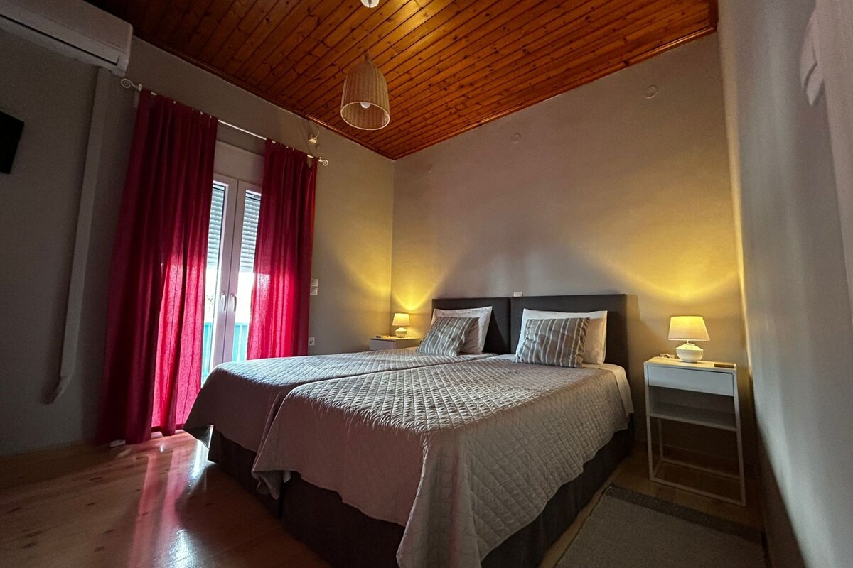A double bed with two bedside tables, a wooden ceiling, and a window with red curtains.