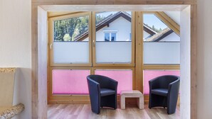 Een kamer met een raam met houten kader, twee zwarte stoelen, een klein houten krukje en een rieten stoel.