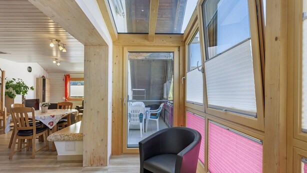 Een kamer met een houten plafond, een eettafel met stoelen, een bankje en een groot raam met jaloezieën.