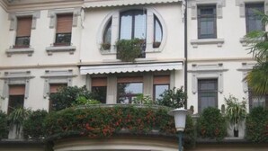 Ein Gebäude im klassischen Stil, mit einem zentralen Eingang mit Säulen, zahlreichen Fenstern mit Fensterläden und einem Balkon mit Pflanzen.