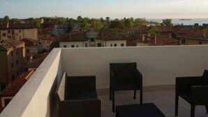 Ein Balkon mit vier dunkel gefärbten Stühlen und einem kleinen Tisch, mit Blick auf eine Küstenstadt, mit Gebäuden und einem weiter entfernten Gewässer.