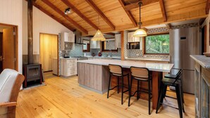 A spacious kitchen with a wooden ceiling, a central island with bar stools, and a built-in stove. - Rincón Nativo 3D y Piscina (Pucón)