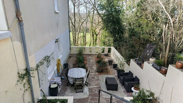 Un escalier menant à un patio aménagé avec une table et des chaises, entouré de plantes en pot et bordé d’un mur blanc.