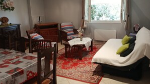 Une pièce comprenant une table à manger, un canapé, une fenêtre avec des rideaux, une petite table sur laquelle est posé un vase, et une chaise.