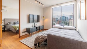 A modern bedroom with a large window, a bed, a TV, and a city view. - Cozy 1-bedroom apartment in peaceful Las Condes with AC (Las Condes)