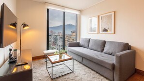 A modern living room with a grey sofa, a glass coffee table, and a large window offering a city view. - Cozy 1-bedroom apartment in peaceful Las Condes with AC (Las Condes)