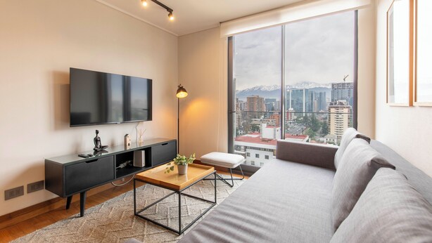 A modern living room with a large window, a flat-screen TV, a sofa, a coffee table, and a small side table. - Cozy 1-bedroom apartment in peaceful Las Condes with AC (Las Condes)