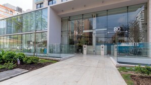 A modern building with large glass windows, numbered doors, and a paved entrance area with greenery. - Cozy 1-bedroom apartment in peaceful Las Condes with AC (Las Condes)