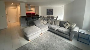 Ein modernes Wohnzimmer mit einem grauen Ecksofa, einem grauen Teppich und einer Küche im Hintergrund.
