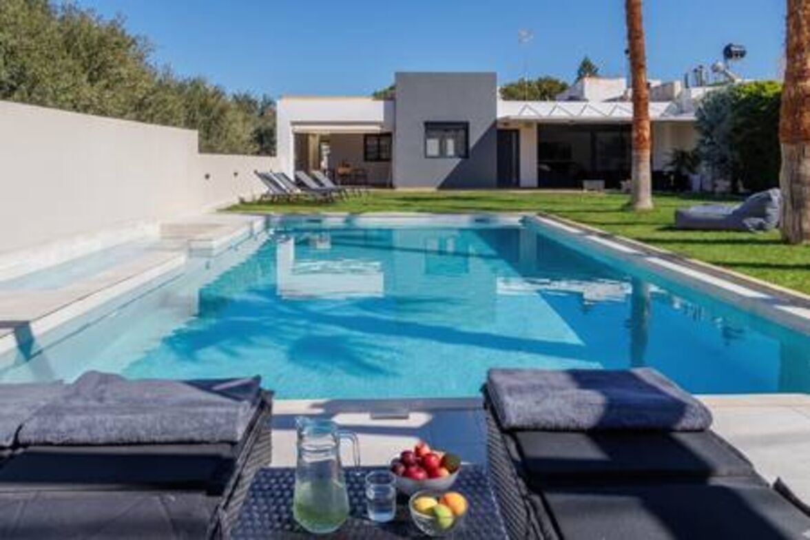 Uma área moderna junto à piscina, com espreguiçadeiras, uma mesa com jarra e frutas, e um edifício contemporâneo ao fundo.