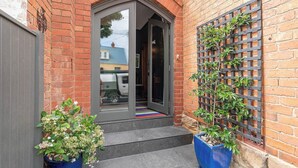 A brick building with a dark-colored entrance door, a small set of steps leading up to it, and two potted plants on either side. - The Church House Spacious Heritage Escape for Two (North Hobart)